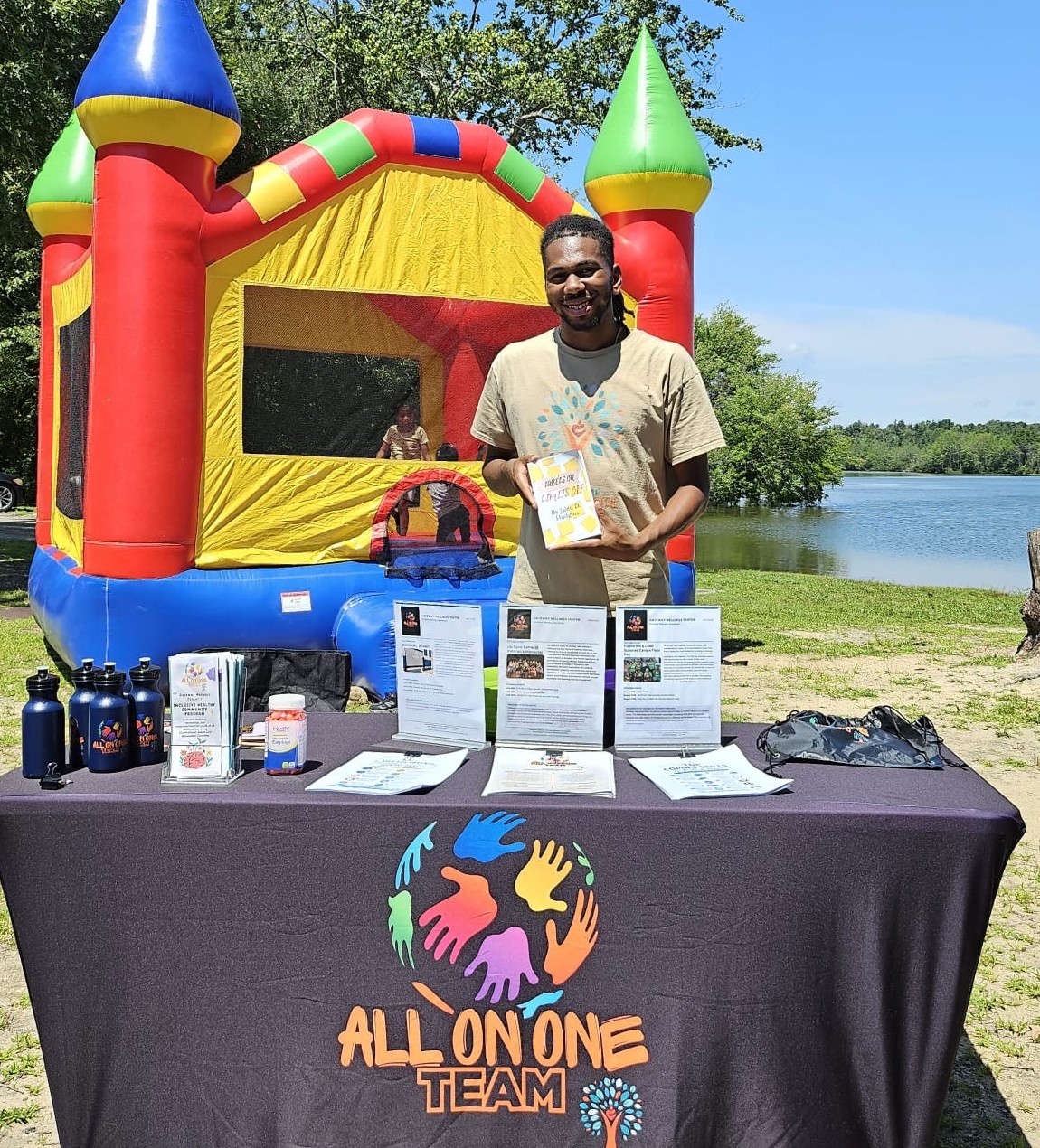 Person holding a book at an outdoor community resource table with informational materials in front of a colorful bounce house near a lakeside park.